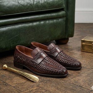 May include: A pair of dark brown leather loafers with woven detailing. The shoes have a classic design with a low heel and a leather strap across the top. A gold-colored shoe horn rests nearby.