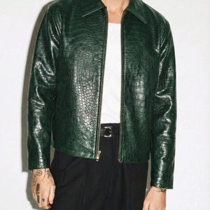 Op de afbeelding: Donkergroene leren jas met ritssluiting en kraag. De jas heeft een textuur die lijkt op krokodillenleer. Gedragen met een wit shirt en zwarte broek.