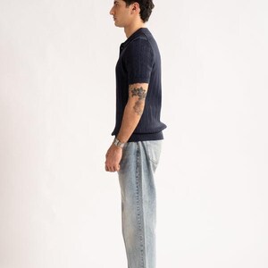 May include: A navy blue polo shirt with a ribbed texture, light blue jeans, and beige shoes. The outfit is styled on a white background. The model is wearing a silver watch and has a tattoo on their arm.