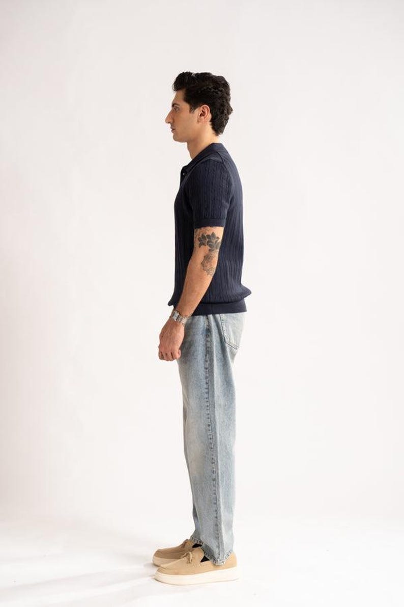 May include: A navy blue polo shirt with a ribbed texture, light blue jeans, and beige shoes. The outfit is styled on a white background. The model is wearing a silver watch and has a tattoo on their arm.