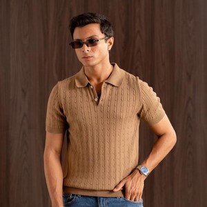 May include: A tan short-sleeved polo shirt with a textured knit design. The shirt has a collared neckline and is worn with blue jeans. The person is wearing sunglasses and a watch.