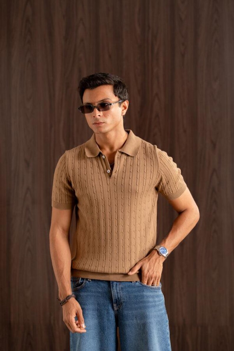 May include: A tan short-sleeved polo shirt with a textured knit design. The shirt has a collared neckline and is worn with blue jeans. The person is wearing sunglasses and a watch.