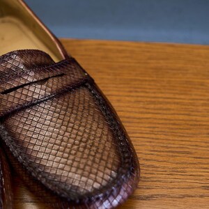 May include: A brown penny loafer shoe featuring a textured, grid-like pattern across the upper surface. The shoe has a classic moccasin-style construction with a horizontal strap across the vamp and visible stitching along the rounded toe edge.