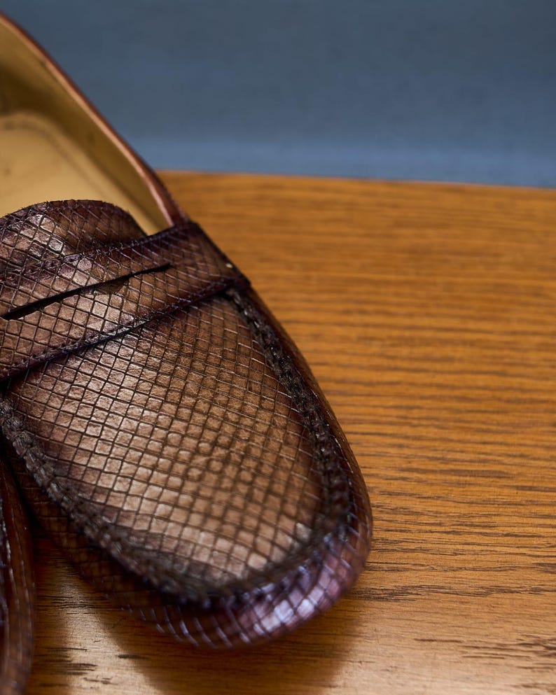 May include: A brown penny loafer shoe featuring a textured, grid-like pattern across the upper surface. The shoe has a classic moccasin-style construction with a horizontal strap across the vamp and visible stitching along the rounded toe edge.