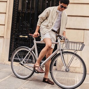 May include: A silver bicycle with a front basket. The bicycle has black tires and is parked on a stone walkway. The person is wearing a light tan jacket, white shorts, and brown sandals.