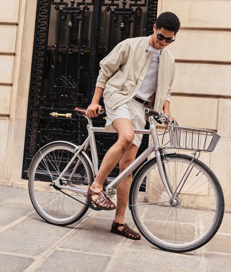 May include: A silver bicycle with a front basket. The bicycle has black tires and is parked on a stone walkway. The person is wearing a light tan jacket, white shorts, and brown sandals.