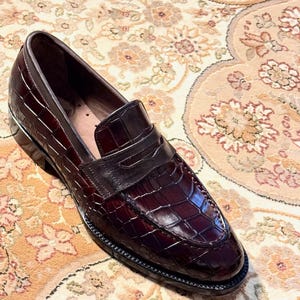 Handcrafted burgundy crocodile embossed leather penny loafers for men featuring a classic apron toe, polished finish, and durable welted sole, displayed on an ornate traditional rug.