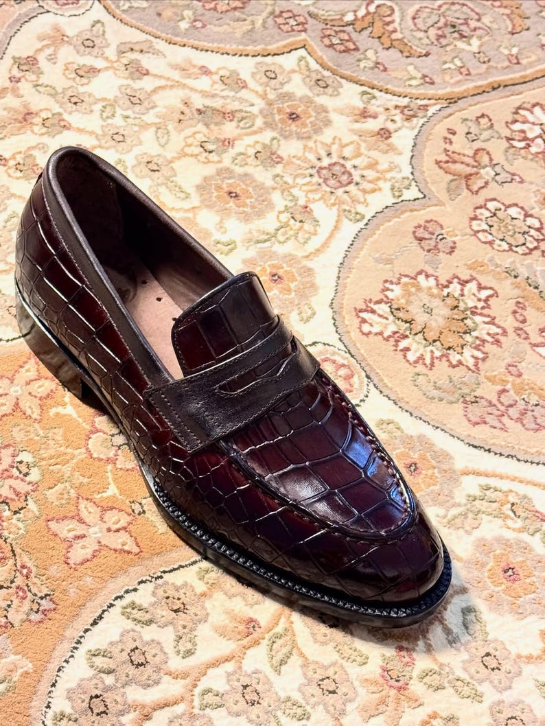 Handcrafted burgundy crocodile embossed leather penny loafers for men featuring a classic apron toe, polished finish, and durable welted sole, displayed on an ornate traditional rug.