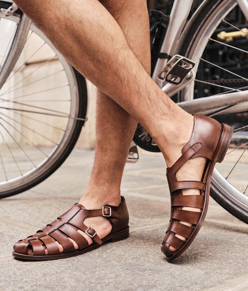 May include: Brown leather sandals with a cage-like design. The sandals have multiple straps and a buckle closure. The shoes are worn by a person near a bicycle. The sandals are a dark brown color.