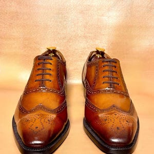 Puede incluir: Un par de zapatos de vestir de cuero marr&oacute;n con intrincados detalles de brogue y un dise&ntilde;o cl&aacute;sico de punta de ala. Los zapatos tienen un acabado pulido, cordones marrones oscuros y una suela negra. Se muestran sobre un fondo neutro.