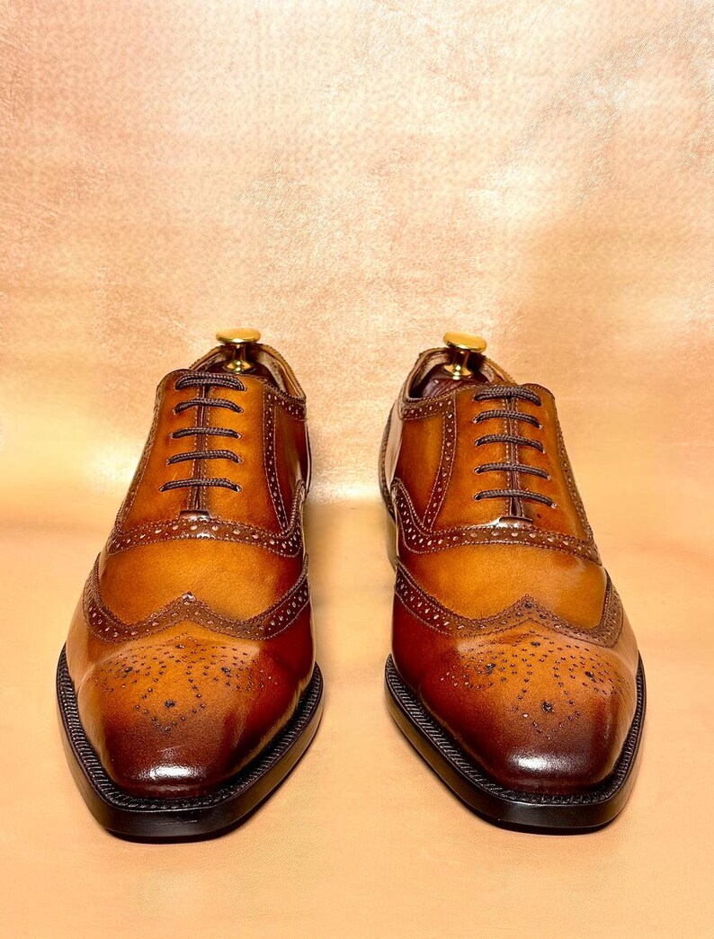 Puede incluir: Un par de zapatos de vestir de cuero marr&oacute;n con intrincados detalles de brogue y un dise&ntilde;o cl&aacute;sico de punta de ala. Los zapatos tienen un acabado pulido, cordones marrones oscuros y una suela negra. Se muestran sobre un fondo neutro.