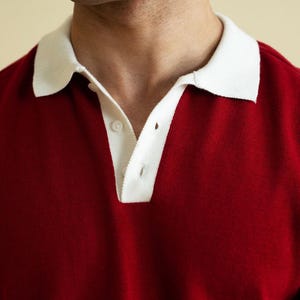 Op de afbeelding: Een close-up van een rode poloshirt met een witte kraag. De shirt heeft een knoopsluiting met drie knopen en is gemaakt van een zacht, gebreid materiaal. De kraag is helderwit, wat contrasteert met het diepe rood van de shirt.