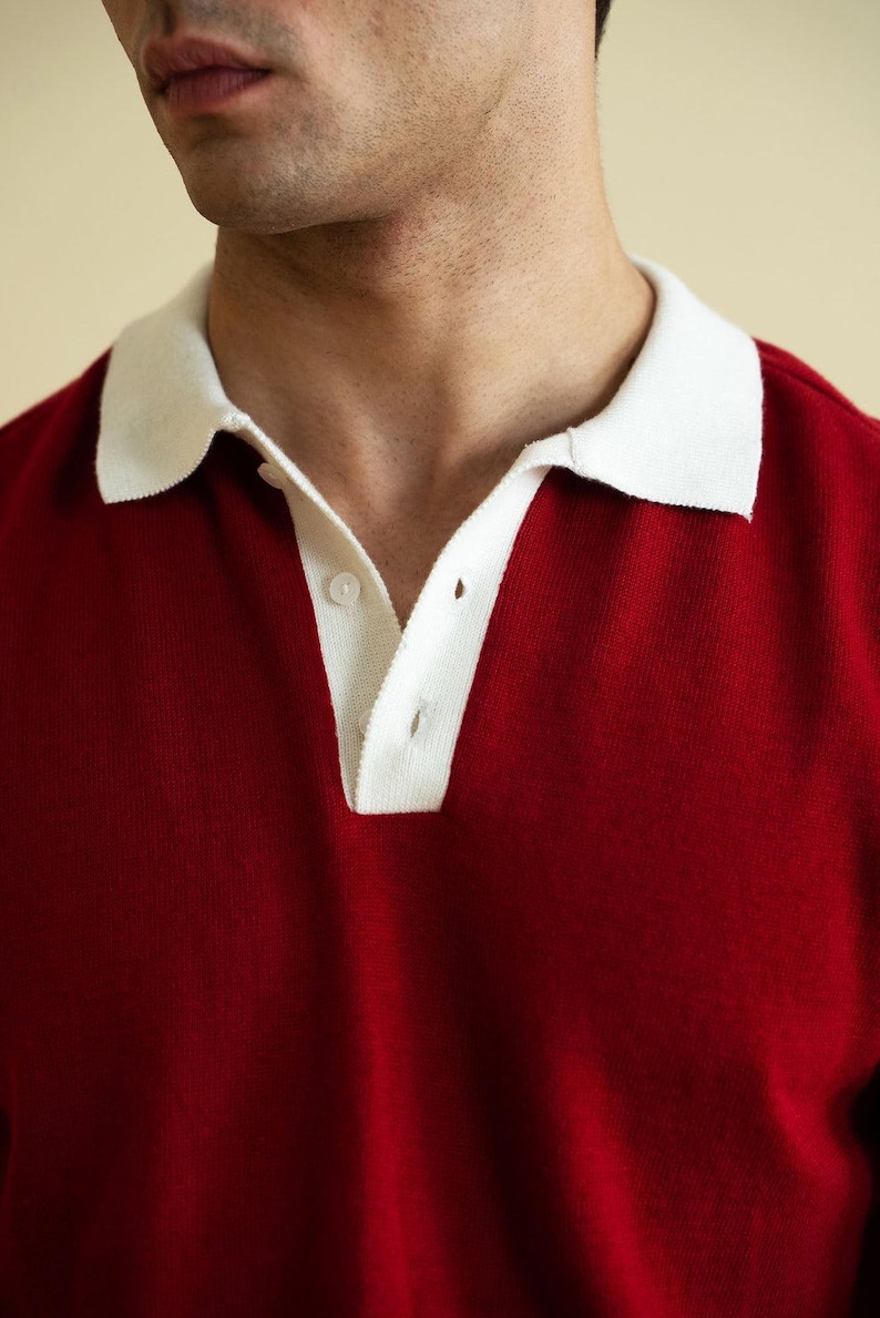 Op de afbeelding: Een close-up van een rode poloshirt met een witte kraag. De shirt heeft een knoopsluiting met drie knopen en is gemaakt van een zacht, gebreid materiaal. De kraag is helderwit, wat contrasteert met het diepe rood van de shirt.