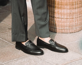 May include: A pair of black leather loafers with silver-tone hardware. The shoes have a classic design with a rounded toe and a slip-on style. The loafers are worn with dark gray trousers.