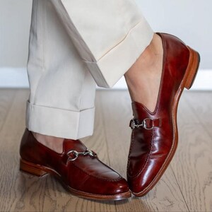 May include: A pair of polished, burgundy leather loafers with silver-tone metal horsebit detailing. The shoes have a classic design with a low heel and are paired with cream-colored trousers. The loafers are on a wooden floor.