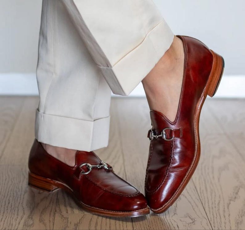 May include: A pair of polished, burgundy leather loafers with silver-tone metal horsebit detailing. The shoes have a classic design with a low heel and are paired with cream-colored trousers. The loafers are on a wooden floor.