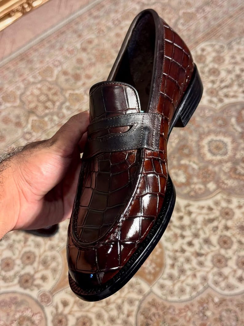 Handcrafted men's penny loafers in dark brown alligator embossed leather, featuring a high-shine polished finish and classic saddle strap detail. Elegant exotic skin slip-on dress shoes for formal wear or weddings.