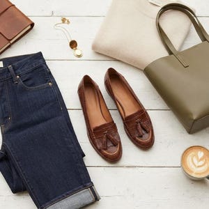 May include: A pair of brown leather tassel loafers with a crocodile-embossed texture, dark blue denim jeans, a beige knit sweater, an olive green tote bag, a gold locket necklace, a brown leather notebook, and a cup of coffee with latte art.