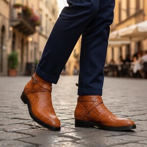 Könnte beinhalten: Hellbraune Leder-Ankle-Boots mit Krokodil-Effekt und Schnallenriemen. Die Stiefel haben eine schwarze Sohle und werden mit marineblauen Hosen getragen. Ein modisches Schuhwerk.