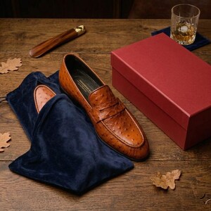 K&ouml;nnte beinhalten: Braune Leder-Loafer mit strukturierter Oberfl&auml;che, pr&auml;sentiert mit einem marineblauen Samt-Schuhbeutel und einer roten Box. Ein h&ouml;lzerner Schuhanzieher und ein Glas bernsteinfarbener Fl&uuml;ssigkeit liegen daneben auf einer Holzoberfl&auml;che.