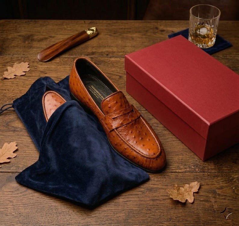 K&ouml;nnte beinhalten: Braune Leder-Loafer mit strukturierter Oberfl&auml;che, pr&auml;sentiert mit einem marineblauen Samt-Schuhbeutel und einer roten Box. Ein h&ouml;lzerner Schuhanzieher und ein Glas bernsteinfarbener Fl&uuml;ssigkeit liegen daneben auf einer Holzoberfl&auml;che.