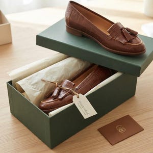 May include: A pair of brown leather loafers with a crocodile-embossed texture, featuring tassels on the vamp. One shoe rests on the lid of a dark green box, while the other is inside, accompanied by a tag labeled Handcrafted and a small card.