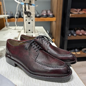 May include: A pair of burgundy leather dress shoes with black laces, displayed on a light-colored surface. The shoes have a textured finish and a classic design. Shelves with other shoes and a sewing machine are in the background.