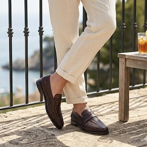 May include: Dark brown woven leather loafers with a rounded toe. The shoes are worn with beige linen pants. A glass of iced tea with a lemon slice sits on a wooden table.