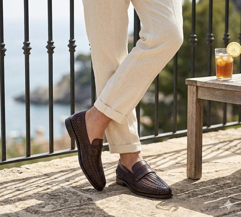 May include: Dark brown woven leather loafers with a rounded toe. The shoes are worn with beige linen pants. A glass of iced tea with a lemon slice sits on a wooden table.