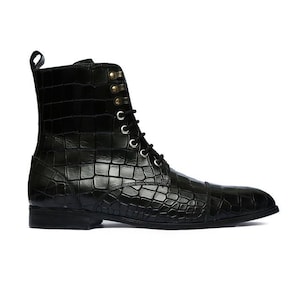 May include: Black leather ankle boot with a crocodile-effect texture. The boot has a lace-up fastening with silver eyelets and a small pull tab. It features a rounded toe and a low heel. The boot is approximately 20 cm high.