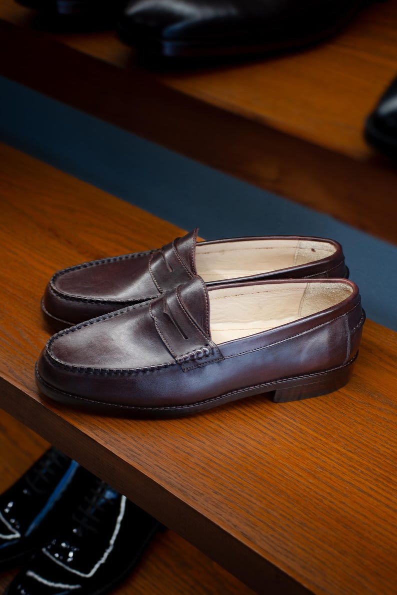 May include: A pair of dark brown leather penny loafers on a wooden shelf. The shoes have a classic design with a rounded toe, a strap across the vamp, and a low heel. The leather appears smooth and polished.