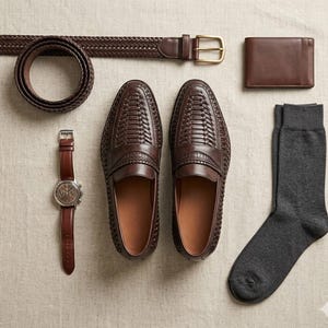 May include: A flat lay of men's accessories in shades of brown and gray. Includes woven leather loafers, a braided belt, a leather wallet, a watch with a brown leather strap, and a pair of gray socks.