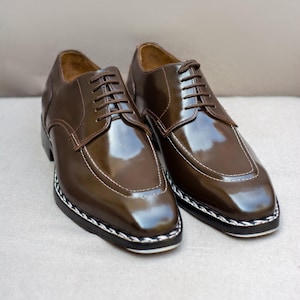 May include: A pair of brown leather dress shoes with a glossy finish. The shoes feature a square toe design, white stitching, and a black and white braided trim around the sole. Brown laces complete the look.