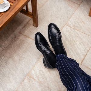 Pu&ograve; includere: Mocassini in pelle nera con finitura strutturata. Le scarpe sono abbinate a pantaloni gessati blu navy. Un tavolo di legno e una tazza di caff&egrave; sono visibili sullo sfondo.