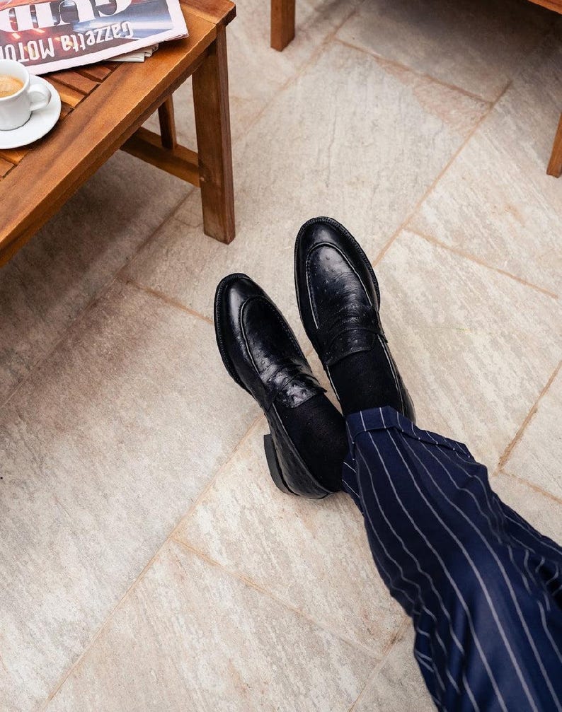 Pu&ograve; includere: Mocassini in pelle nera con finitura strutturata. Le scarpe sono abbinate a pantaloni gessati blu navy. Un tavolo di legno e una tazza di caff&egrave; sono visibili sullo sfondo.