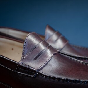 May include: Close-up of a pair of brown leather penny loafers. The shoes have a classic design with a stitched vamp and a strap across the top. The leather has a polished finish, and the shoes are set against a blue background.