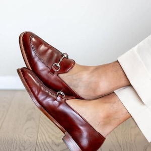 May include: A pair of brown leather loafers with silver-colored metal accents. The shoes have a classic design with a rounded toe and a low heel. The loafers are shown on a wooden floor.