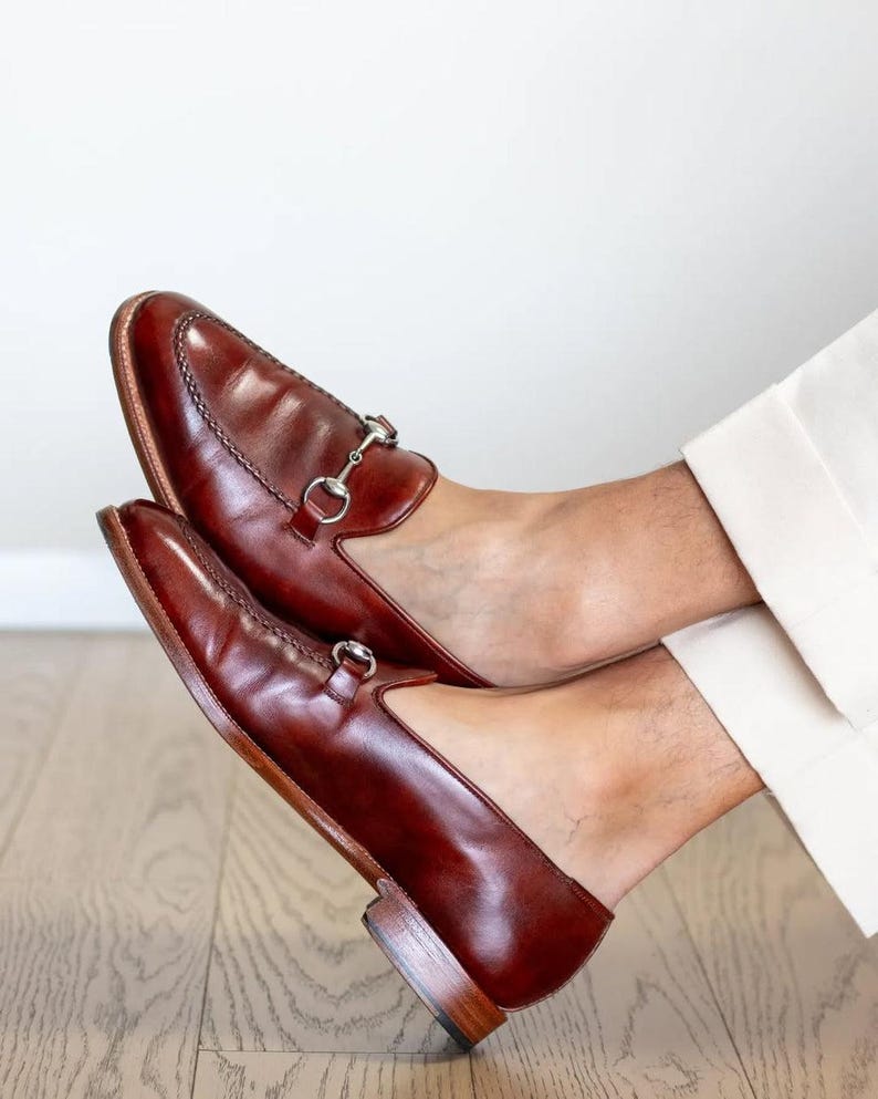 May include: A pair of brown leather loafers with silver-colored metal accents. The shoes have a classic design with a rounded toe and a low heel. The loafers are shown on a wooden floor.