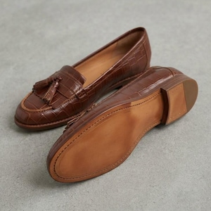 May include: A pair of brown loafers with a crocodile-embossed texture, featuring a tassel detail on the top of one shoe and a smooth, light brown sole on the other. The shoes have a low heel and a classic slip-on design with visible stitching.