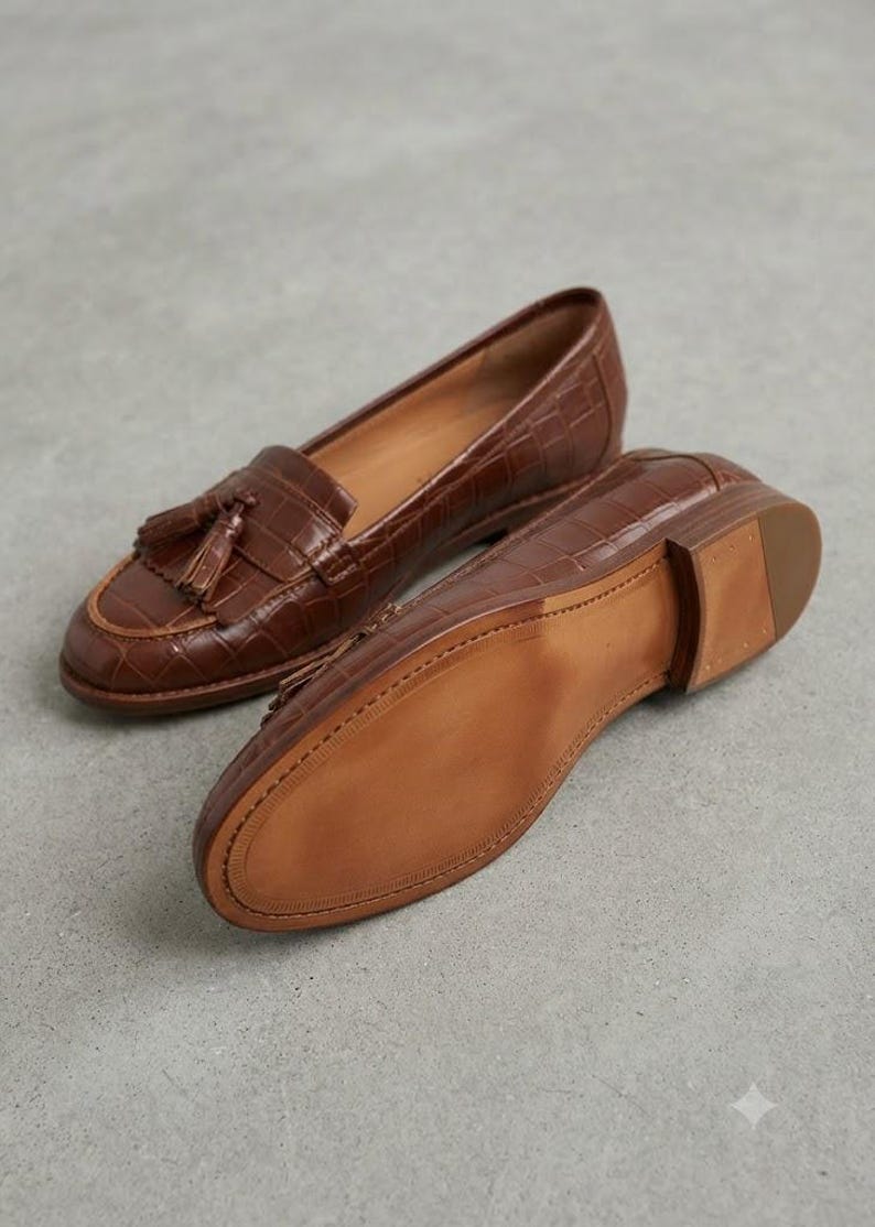 May include: A pair of brown loafers with a crocodile-embossed texture, featuring a tassel detail on the top of one shoe and a smooth, light brown sole on the other. The shoes have a low heel and a classic slip-on design with visible stitching.