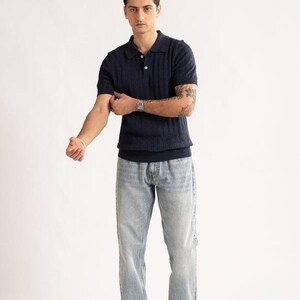 May include: A navy blue polo shirt with a textured pattern and short sleeves. The shirt is paired with light blue jeans and beige loafers. The model is wearing a silver watch and a bracelet.