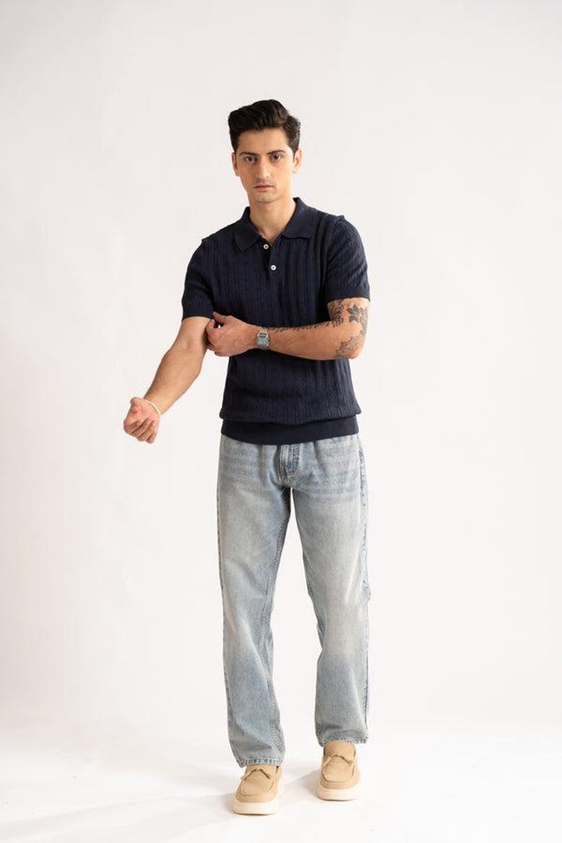 May include: A navy blue polo shirt with a textured pattern and short sleeves. The shirt is paired with light blue jeans and beige loafers. The model is wearing a silver watch and a bracelet.