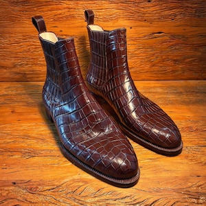 Peut inclure: Une paire de bottines en cuir marron avec une texture peau de crocodile. Les bottines ont un bout pointu et une tirette en haut. Les bottines sont présentées sur une surface en bois.