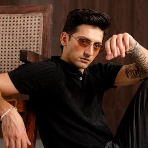 May include: A black polo shirt with a textured knit design. The model is wearing brown-tinted oval sunglasses. The person is sitting on a wooden chair with a woven back. The person is also wearing a bracelet.