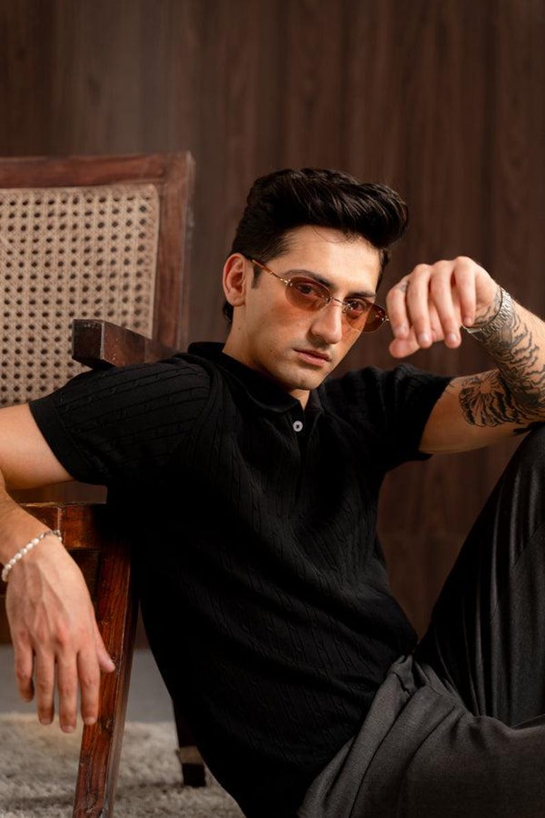 May include: A black polo shirt with a textured knit design. The model is wearing brown-tinted oval sunglasses. The person is sitting on a wooden chair with a woven back. The person is also wearing a bracelet.