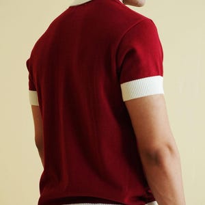 Op de afbeelding: Een bordeauxrood shirt met korte mouwen en witte randen op de kraag en mouwen. Het shirt is gemaakt van een zachte gebreide stof. Het model draagt een lichtgeleurde broek. Het shirt heeft een klassieke, retro stijl.