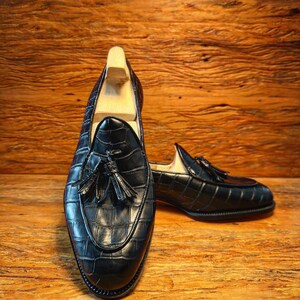 Puede incluir: Mocasines de cuero negro con textura de cocodrilo y detalles de borlas. Los zapatos se exhiben sobre una superficie de madera. El dise&ntilde;o presenta una puntera redondeada y un tac&oacute;n bajo. Adecuados para uso formal o informal.