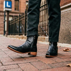 Könnte beinhalten: Schwarze Leder-Ankle Boots mit Schlangenmuster. Die Stiefel haben einen seitlichen Reißverschluss, eine spitze Zehenpartie und einen niedrigen Holzabsatz. Getragen mit dunkelgrauen Jeans.