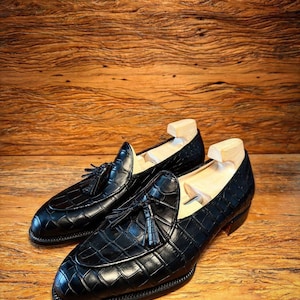 Puede incluir: Un par de mocasines de cuero negro con un patr&oacute;n de cocodrilo y detalles de borlas. Los zapatos se exhiben sobre una superficie de madera, mostrando su acabado pulido y dise&ntilde;o cl&aacute;sico. Se insertan hormas.