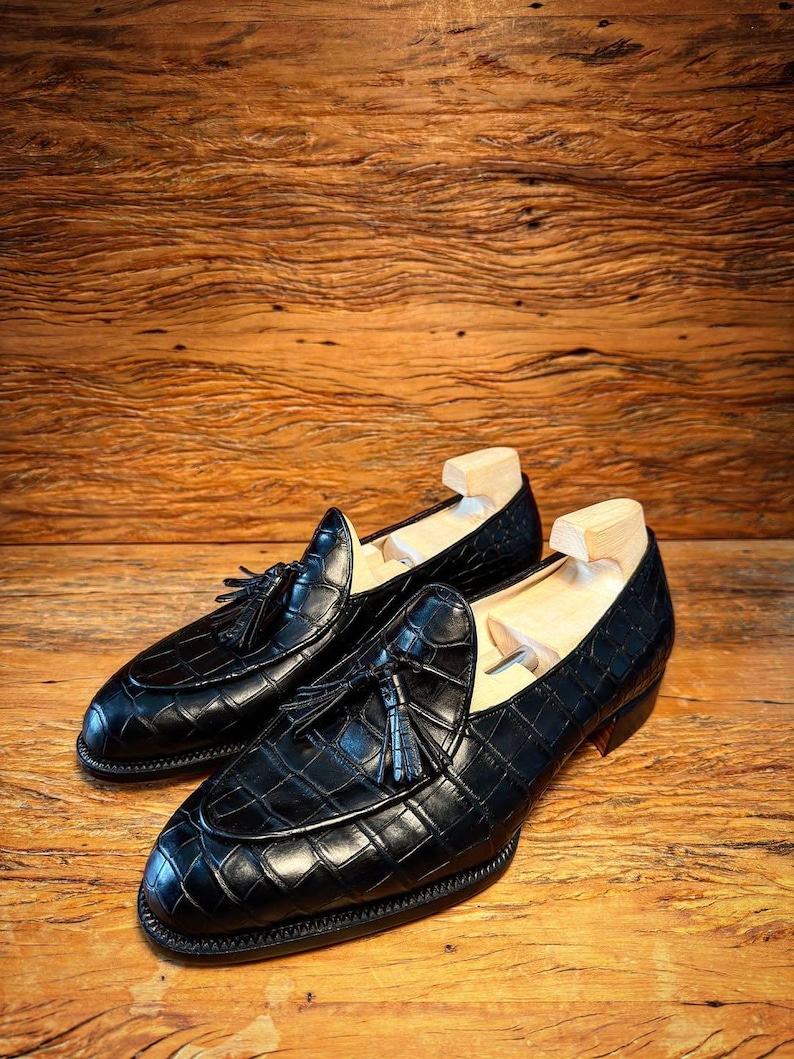 Puede incluir: Un par de mocasines de cuero negro con un patr&oacute;n de cocodrilo y detalles de borlas. Los zapatos se exhiben sobre una superficie de madera, mostrando su acabado pulido y dise&ntilde;o cl&aacute;sico. Se insertan hormas.